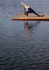 one alone young latina hispanic girl woman doing extended side extended angle yoga pose Parsvakonasana on pier of lake pond river Reflection on water surface.