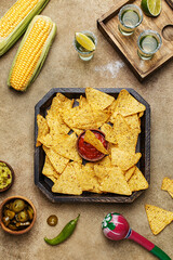 Nachos corn chips with guacamole and salsa dressing, fresh corn cobs, marinated jalapeño, chili pepper and tequila shots on beige concrete background. Mexican party snack. Top view 