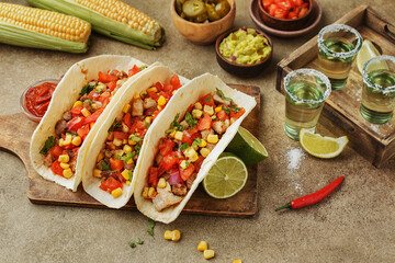 Tacos with chicken, tomatoes and corn with guacamole and salsa sauce, lime, fresh corn cobs, chili peppers and tequila shots on beige concrete background. Mexican party snacks