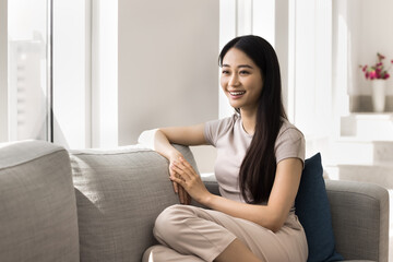 Obraz premium Asian woman sits on modern sofa in sunlit living room