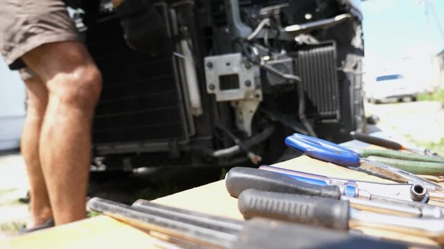 Male hand of mechanic in glove taking screwdriver from wooden plank surface for repair radiator of car outdoor. Professional repairman making maintenance of auto at workplace. Focus on foreground