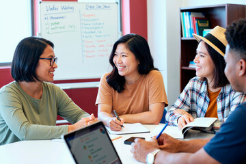 Group of young Asian women and young Black man sitting at table discussing ideas and writing in notebooks during meeting in modern office setting with whiteboard in background
