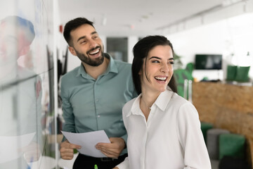 Overjoyed millennial man woman coworkers laughing near visual schedule board