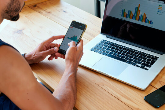Young man analyzing financial data on smartphone while sitting at desk with open laptop displaying colorful bar charts, focusing on business analytics