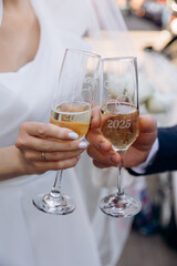 Close-up of a newlywed couple's hands clinking personalized champagne flutes engraved with '2025' and their names during a wedding celebration