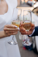 Newlywed couple toasting with personalized champagne glasses engraved with '2025' and their names, with a white wedding car visible in the background