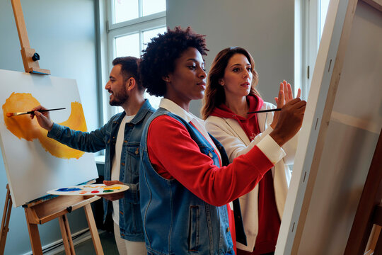 Multiethnic group of young adults painting on canvases in art studio, Black woman and man using brushes while woman observing and discussing creative process