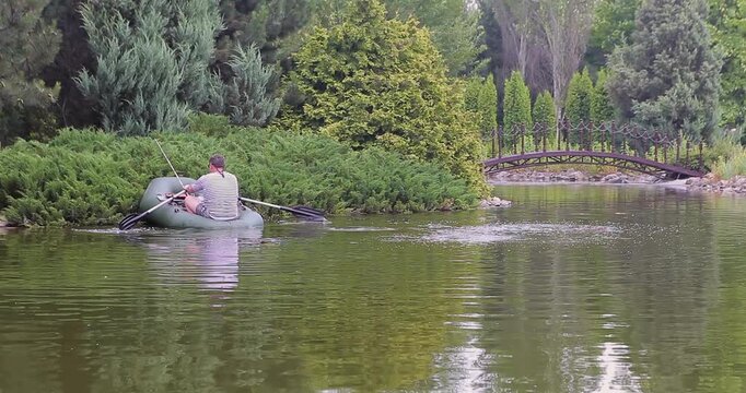 A man sails a rubber boat across an artificial pond. A man fishes in a small rubber boat in a private ornamental pond. A beautiful summer landscape on the water.