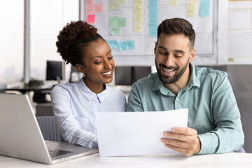 Smiling two diverse young businesspeople reading papers at workplace desk