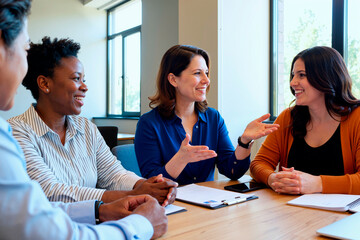 Group of middle aged women and Black woman sitting at table engaging in discussion, smiling and gesturing while collaborating in modern office setting during meeting
