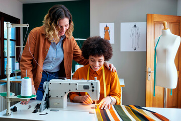 Young woman standing beside young Black woman operating sewing machine, both smiling and collaborating in fashion design studio with dress sketches visible