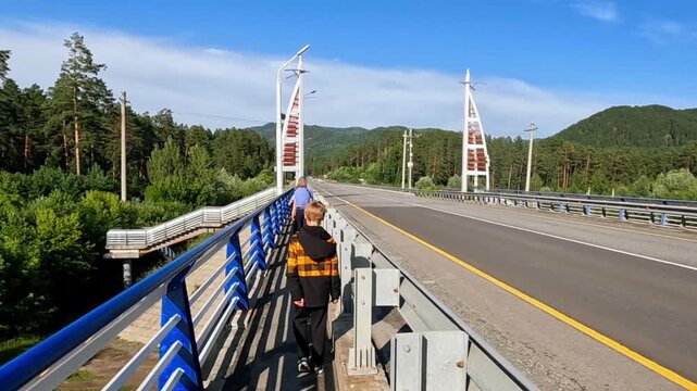 A boy in a jacket walks along the pedestrian section of a modern bridge against a backdrop of green mountains and a pine forest. Summer trip, road and architecture in nature.
