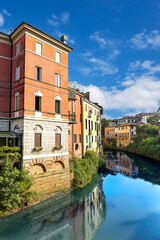 The Baciglione River gently flows through Vicenza, surrounded by colorful historic buildings. A bright blue sky reflects in the calm waters, creating a peaceful scene.