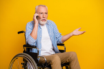 Funny grandpa on a wheelchair talks on his phone and gestures with an open hand against a bright yellow backdrop for a warm accessible health and family moment
