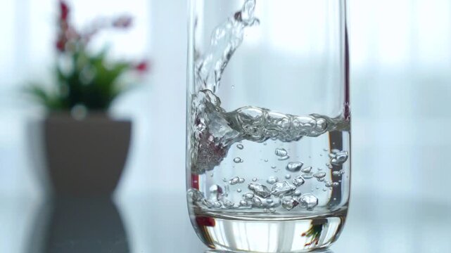 Crystal clear water filling a transparent glass on a table, with splashes and bubbles forming in slow motion. Healthy hydration and refreshment concept with a blurred plant in the background