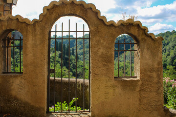 Antica facciata ammalorata di ingresso giardino con cancello ‌e finestra con grate in ferro e cielo con nubi a Pitigliano