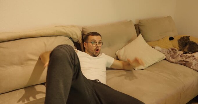 A man relaxes comfortably on the couch when something surprising happens on TV, causing him to sit up in shock and react instantly.