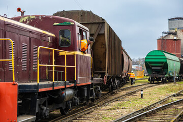 Two railway workers in orange safety jackets coordinate around a maroon diesel locomotive pulling freight wagons on weathered tracks in an industrial rail yard