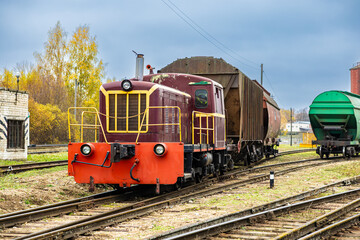 Fototapeta premium A maroon and red diesel locomotive hauls rusty and green freight wagons through a rail yard on an overcast autumn day, surrounded by yellow foliage and tracks