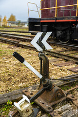 Manual railway switch lever with a black and white directional arrow in the foreground, with train tracks and a freight wagon visible in the distance under cloudy skies