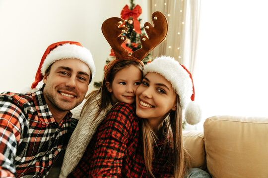 Overjoyed parents with daughter in reindeer antlers, mother and father wearing Santa hats, sitting by the decorated Christmas tree, enjoying family love, warmth, and festive holiday atmosphere.