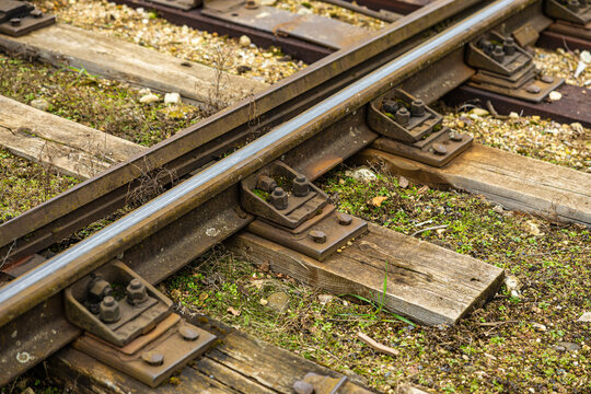 Close-up of an old railway track section with rusty bolts, moss-covered fasteners, and weathered wooden ties surrounded by gravel and green vegetation
