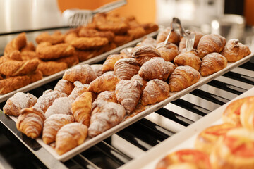 A selection of various baked pastries is arranged neatly on a tray in a bakery. The warm croissants with powdered sugar are highlighted, enticing customers