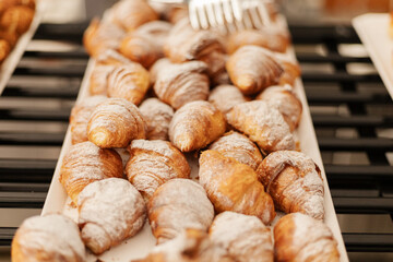 A variety of golden-brown croissants dusted with powdered sugar are arranged neatly on a tray. The...