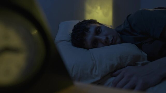 Man lying awake at night, blurry clock silhouette in foreground, warm yellow window light, insomnia night scene