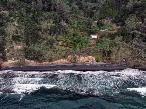 Aerial view of the dark, volcanic sand meeting the turquoise waves at the shore below the vibrant green cliffs of Praia da Amora, Ponta GarÃ§a, Azores, Portugal.