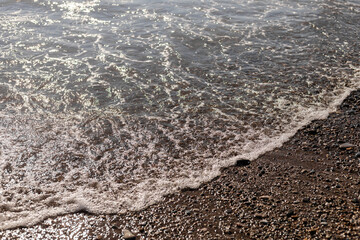 Waves gently roll onto a sandy beach, glistening in the sunlight. Small pebbles dot the shoreline, creating a serene and calming atmosphere by the water's edge