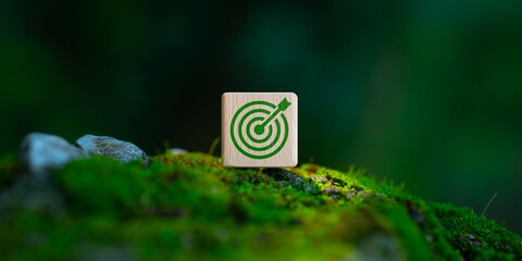 A wooden cube with a green target icon sits on a mossy ground amidst lush nature, symbolizing sustainability goals, environmental protection, eco-friendly objectives and achieving green goals.