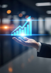A human male hand in the lower frame, cupped upward, holding a glowing blue financial graph. The upward arrow and rising bar charts symbolize business success and economic growth. Background shows a b