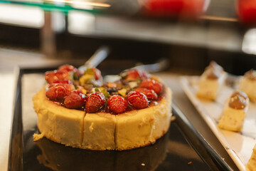 A cheesecake sits elegantly on a black tray, adorned with glossy strawberries and a mix of vibrant fruits. The setting is a dessert display in a bright venue, inviting guests