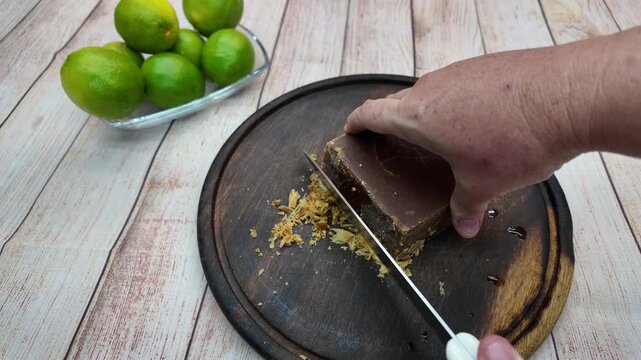 Hand cutting panela with knife on wooden board beside fresh limes