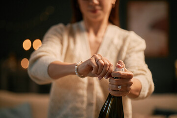 Caucasian young adult woman opening champagne bottle with both hands, standing indoors with blurred background, focus on hands and bottle, face partially visible