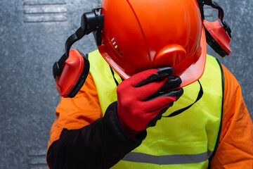 Orange protective safety helmet with ear muffs held by a worker wearing a reflective vest. Concept...