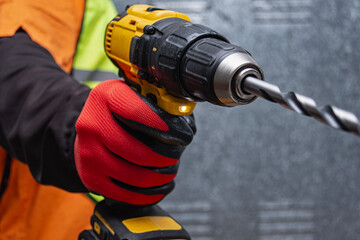 Close-up of a worker holding a yellow cordless drill with a drill bit, wearing protective gloves and safety vest. Concept of construction, power tools, and professional craftsmanship.
