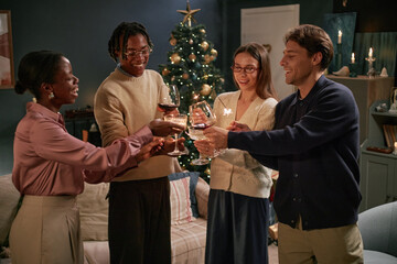 Group of diverse young adults friends standing together clinking wine glasses in front of decorated Christmas tree, celebrating holiday gathering