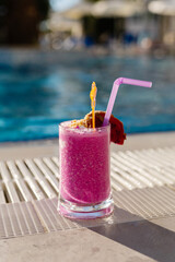 A colorful pink smoothie sits on a sunlit pool deck, surrounded by a serene water backdrop. Fruit garnishes enhance its tropical appeal on a warm afternoon