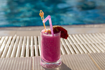 A vibrant pink cocktail sits on the edge of a sunlit pool. The drink is garnished with a slice of fruit and a playful straw, offering a perfect refreshment for a warm day