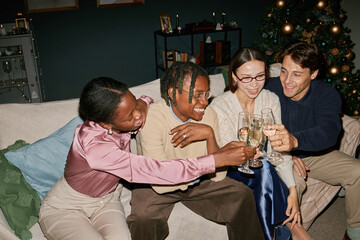 Diverse group of young adults sitting on sofa clinking champagne glasses, celebrating together near...