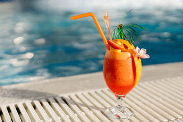 Bright orange cocktail garnished with fruit and decorative elements rests on a poolside platform. Sunlight reflects off water, creating a cheerful summer atmosphere