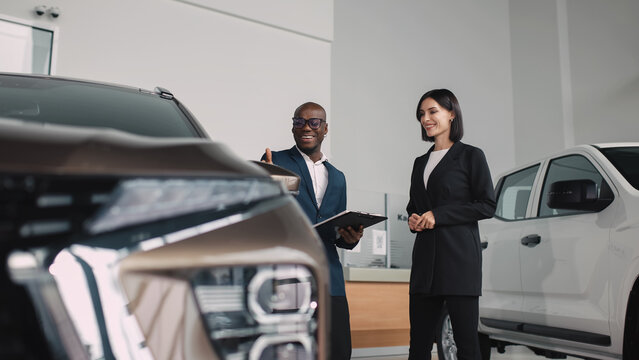 African American man in a suit presenting a car to a woman in a blazer, showcasing features and benefits in a modern dealership environment with bright lighting