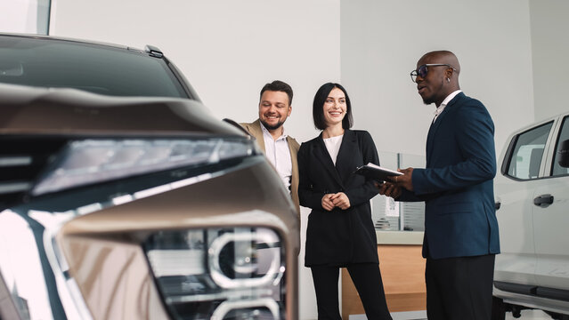 African American man in a suit discussing car features with a couple in a dealership, showcasing the car buying experience and customer service interaction
