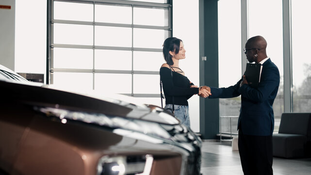 Woman shaking hands with a car salesman in a modern dealership, showcasing a sleek vehicle in the foreground, emphasizing the car buying experience and customer satisfaction