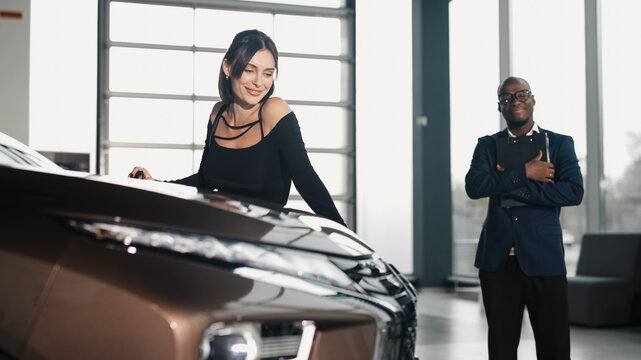 Smiling woman examining a sleek brown car in a modern dealership, with a man in a suit observing, showcasing the excitement of buying a new vehicle with copy space