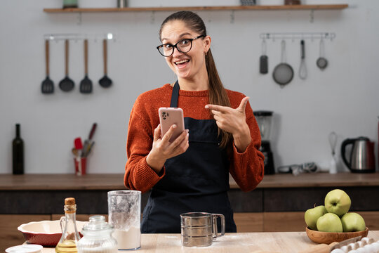 Portrait of woman wearing apron cooking apple pie enjoy making clean food pointing finger on smartphone in kitchen at home. baking, cookies and dessert.