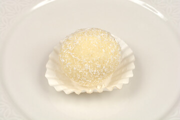 A single, round coconut truffle, lightly dusted with flakes, is centered in a white paper liner on a matching white decorative plate