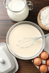 Liquid dough in bowl, whisk and ingredients on wooden table, flat lay